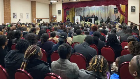 Many people can be seen sat on red chairs looking at a stage where Panashe's funeral was taking place.