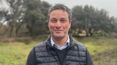 A man smiling wearing a thin puffer vest on top of a black jumper and plaid blue shirt. Behind is blurred greenery and trees with grey skies behind.