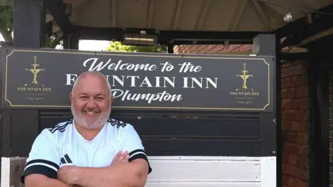 A man stands with his arms folded outside a pub. He is bald and wearing a white T-shirt. He is smiling. 