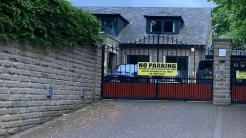 LDRS A house pictured from behind some black iron gates. A yellow 'no parking' sign is on the gates.