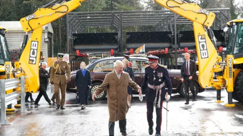 Reuters King Charles III, wearing a long brown coat, and is contingent walk underneath an arch created by the arms of two JCB vehicles. A dark red saloon car can be seen in the background.