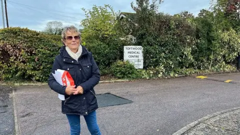 Valerie Savory standing in front of a sign saying Toftwood Medical Centre. She is wearing sunglasses, carrying a bag of prescription medication and wearing a blue coat and jeans.