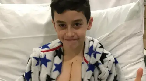 Family photo A boy with dark hair in a dressing gown in a hospital bed with a scar down his chest 