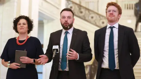 Getty Images Three people standing in front of microphone. A woman on the left with short brown curly hair wearing a black dress an an orange necklace. A brown haired man wearing a suit with a teal tie in the middle and a ginger man wearing a black suit with a navy tie on the right.