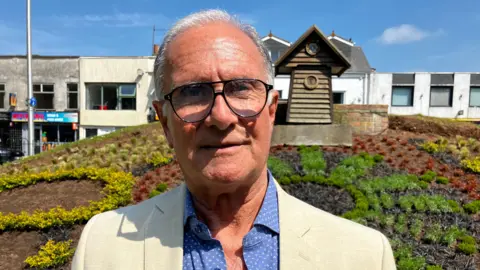 Councillor Mike Solomon standing in front of a wooden clock and plant display in a town centre. He is wearing large black glasses, a beige jacket and blue shirt.