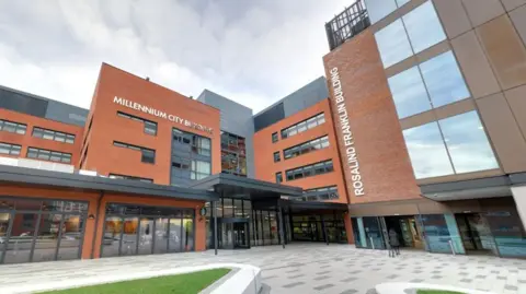 The outside of the University of Wolverhampton's Millennium City and Rosalind Franklin Buildings, both modern multi-storey buildings with glass windows at street level. A grey concrete square with patches of landscaped grass lies outside.