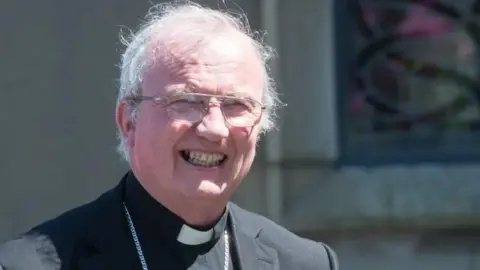 Pacemaker Donal McKeown is smiling at the camera. He is wearing a black jacket white and clerical collar with a large silver chain around his neck. He has white hair and gold glasses