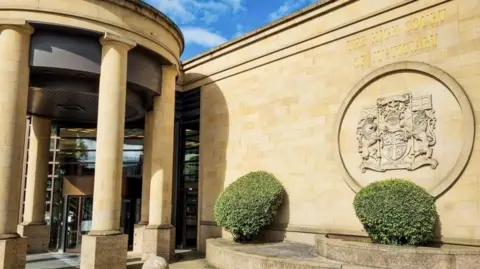 Exterior photograph of the front and entrance to the High Court in Glasgow. The sand-coloured building features a large stone-carved coat of arms above one of two hedges to the right of the entrance, which is a glass panelled revolving door.