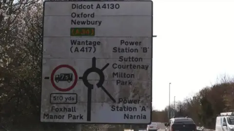 A large road sign appearing before a roundabout. "Narnia" is one of the locations that appears on the sign. Traffic can be seen passing to the right.