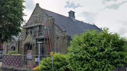 A church with scaffolding on the exterior. It is set back from the street and surrounded by a low, stone wall and shrubbery.
