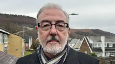 A white man with short grey hair and glasses standing in front of a housing beneath a mountain