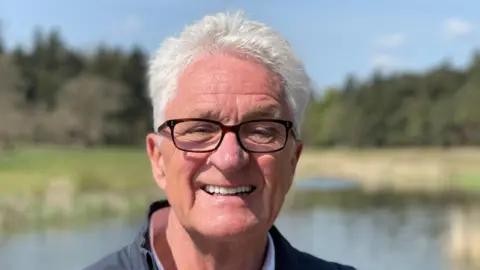 Melvin Benn smiles at the camera on the site of the Latitude Festival. He has white hair and wears glasses and a dark jacket. It is just his face in the shot. Behind him, there is a lake and some trees. The background is out of focus.