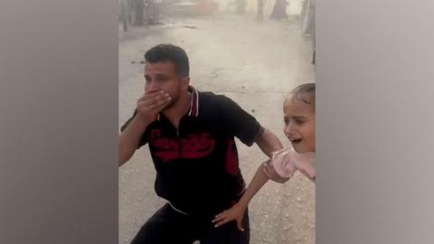 Man and child in shock after Gaza blast, man has hand over mouth and other hand holding girls arm, they are outside on a street with dust from blast and people in the background