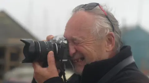 Stephen Squirrell looks into the viewfinder of his camera as he points it to the left. His eye closest to the camera is shut closed as he takes the picture. He has sunglasses on his head and has grey hair, and wears a black jumper. 