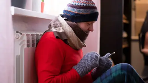 Getty Images A man leaning against a radiator wears a red jumper, a brown and white scarf, a striped multicoloured hat and blue woollen mittens while using his phone. 