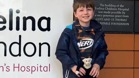 Rufus Quinn is seen smiling while standing next to the Evelina Hospital sign holding a stuffed toy monkey.