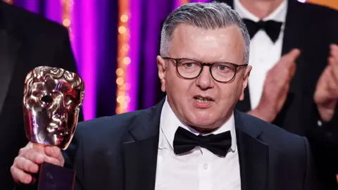 Richard Frediani in a dinner jacket and bow tie holding up a Bafta award with one hand and delivering a speech at the Bafta TV Awards on 11 May