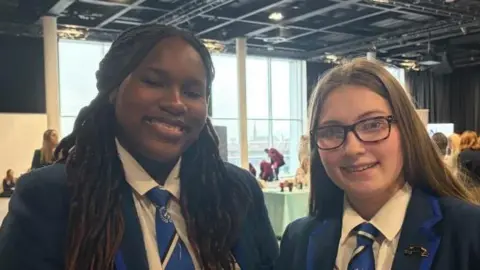 Kayla and Brooke and smiling at the camera. Brooke wears glasses. They are wearing navy school blazers that have lighter blue lining. Their school ties are light blue to match.