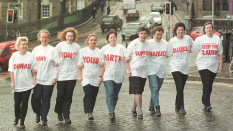 A group of mothers from Perth walk in one long line  to the entrance of the Scottish Parliament - they each wear a white t-shirt with red words which all spell out "Donald, why don't your party support family values, keep the clause".