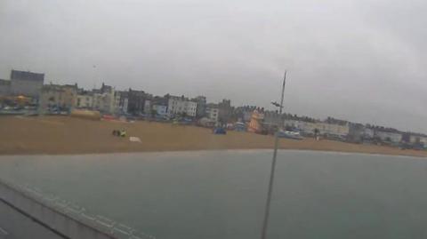 CCTV screenshot of Weymouth beach from the pavilion shows the sea and sandy beach with flat buildings lining the street. On the sand you can see a white cordon and at least three people, two wearing all black and on in a high-vis yellow jacket