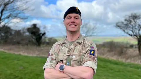 Lieutenant Tim Gilbert stood with his arms crossed and smiling at the camera. He's wearing a beret and camouflage uniform. 