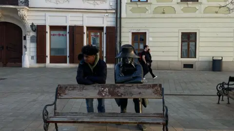 Luka Chijutomi-Ghosh Luka Chijutomi-Ghosh berdiri di belakang bangku di sebelah patung, di Bratislava, mereka berdua memegang pose, bersandar di belakang bangku dengan tangan bersilang.