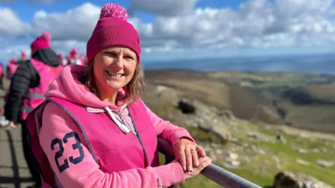 Diane Taylor smiling in front of the rolling Manx hills on a sunny day wearing a pink jumper and bobble hat.