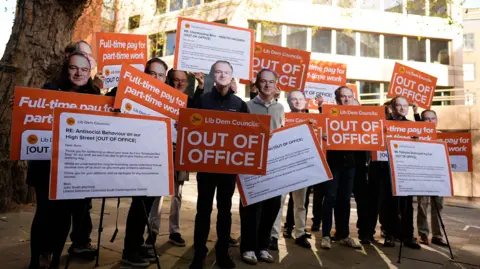 Aaron Chown/PA Wire A group of people wearing cardboard masks bearing Liberal Democrat leader Sir Ed Davey's face and holding up red and white posters that say "Lib Dem Councils: Out of Office" and "Full-time pat for part-time work".