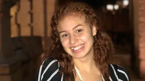 Collect/PA Real Life A smiling woman with curly ginger hair and a black and white striped top in a dark room