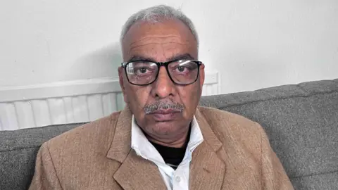 Kate Bradbrook/BBC Close-up of a man wearing glasses and a tan jacket, seated on a sofa indoors.