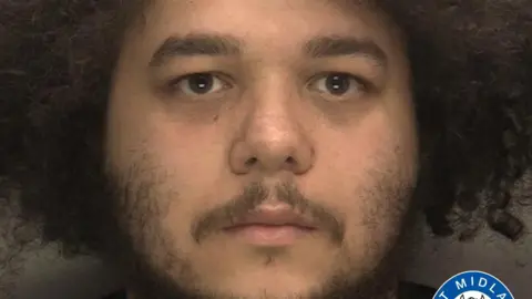 A mugshot of a man with black afro hair and a long black moustache 
