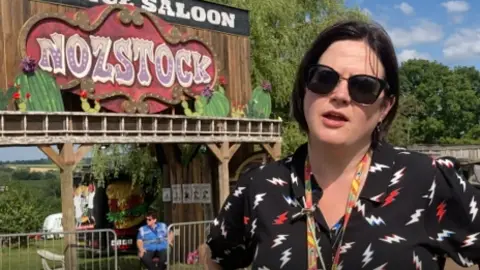 Ms Nosworthy is wearing a patterned top and sunglasses. She has short dark hair and is stood by a entrance to her festival.