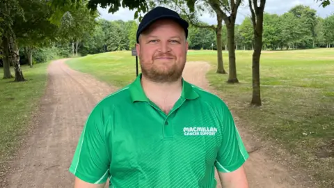 A man stands under trees on a dirt track on a golf course. He is wearing a bright green Macmillan Cancer Support polo shirt and a black cap. He has a short brown beard and he is smiling.