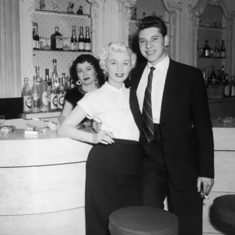 Hulton Archive/Getty Images Two women and a man, dressed smartly, standing at a nightclub bar with bottles of alcohol visible in the background. It is a black and white photo and they are all smiling and looking at the camera.