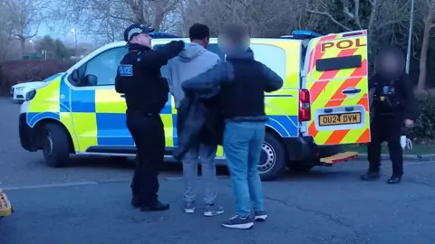 A large police van with police officer standing next to two members of the public. There is another police officer standing next to the back of the van with the door open 