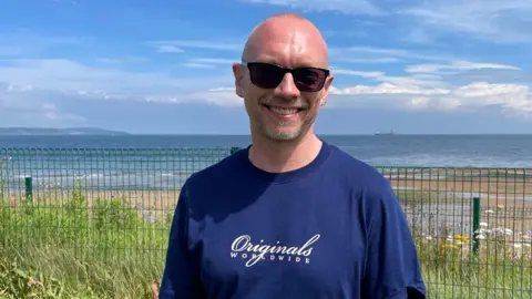 BBC A man wearing a navy t-shirt and sunglasses. He is standing in front of a beach and is smiling at the camera.