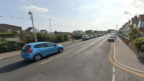 Google A Google Maps image of a junction. There is one blue car driving on the road, and others parked up on either side. 