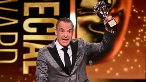 Getty Images Martin Lewis speaking on stage and holding a Bafta aloft in one hand