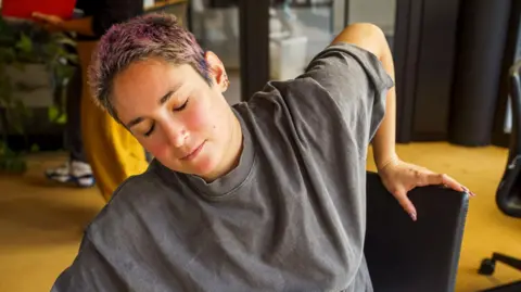 Getty Images Person with cropped pink-dyed hair is leaning to the side in an office chair. They have their eyes closed and an expression of mild discomfort or fatigue on their face.