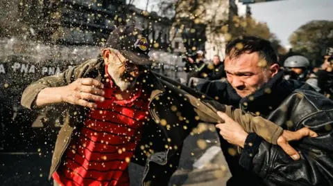 EPA A demonstrator with a white beard and wearing glasses and a baseball cap is being grabbed by a police officer wearing a black leather jacket. The demonstrator is clutching at the officer's sleave during the protest outside Congress in Buenos Aires on 11 September 2024.