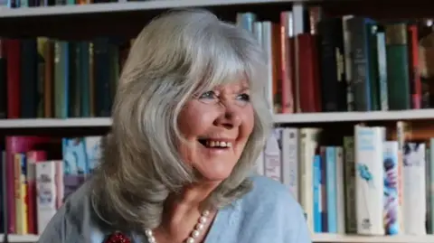Antony Thompson/Thousand Word Media Ltd Image of Dame Jilly Cooper smiling off to the side of the image, wearing a light blue jumper, pearls and has a big smile on her face. A book case can be seen behind her.
