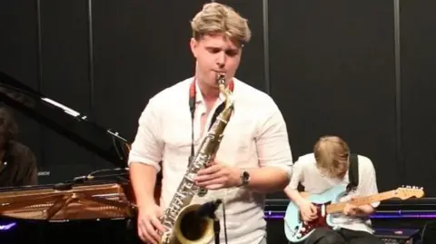 Conor Parker-Delves, playing the saxophone which was stolen in a concert. He has short floppy brown hair and is wearing a white shirt with his sleeves rolled up. In the background is a bass guitar player sitting on a chair, and a person playing a grand piano.