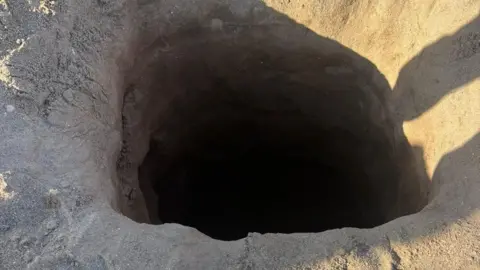 A large, deep hole dug into sandy ground on a beach, with steep, uneven sides and a dark interior where the bottom is not visible. Strong sunlight casts shadows across part of the hole and adjacent sand.
