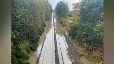 Network Rail Wales & Borders Train tracks are visible poking out of pools of water. Trees line each side