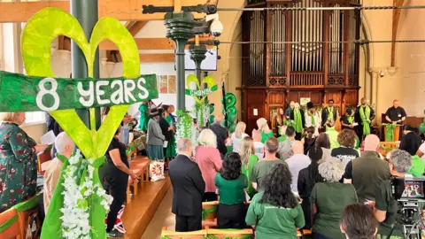 Notting Hill Methodist Church, Lancaster Road. People are sat in wooden pews and the columns are decorated in green and white flowers. 