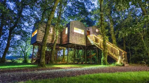 Faraway Treehouses A large tree house sits on stilts amongst trees. The lights inside the treehouse glow and light up the building which is in a forest in Cumbria. 