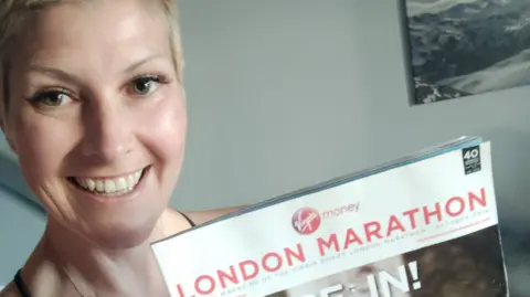 A smiley woman with closely cropped blonde hair holds up a magazine with London Marathon printed on the front 