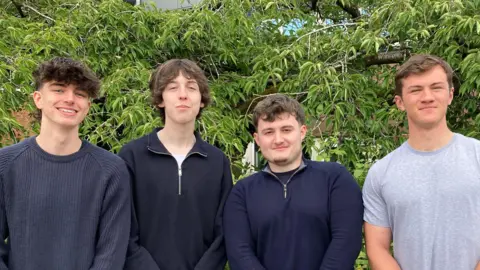 BBC Four young men stand posing at the camera in front of a leafy green tree
