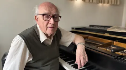 Paul Chrimes sitting at a grand piano wit his arm leaning on the top of it. He is balding with white hair and wearing glasses, a watch, a tank top and a short-sleeved white shirt.