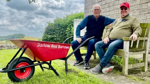 Two men sit on a bench beside a red wheelbarrow. The barrow says “Barlow Red Barrows” printed in white letters.
